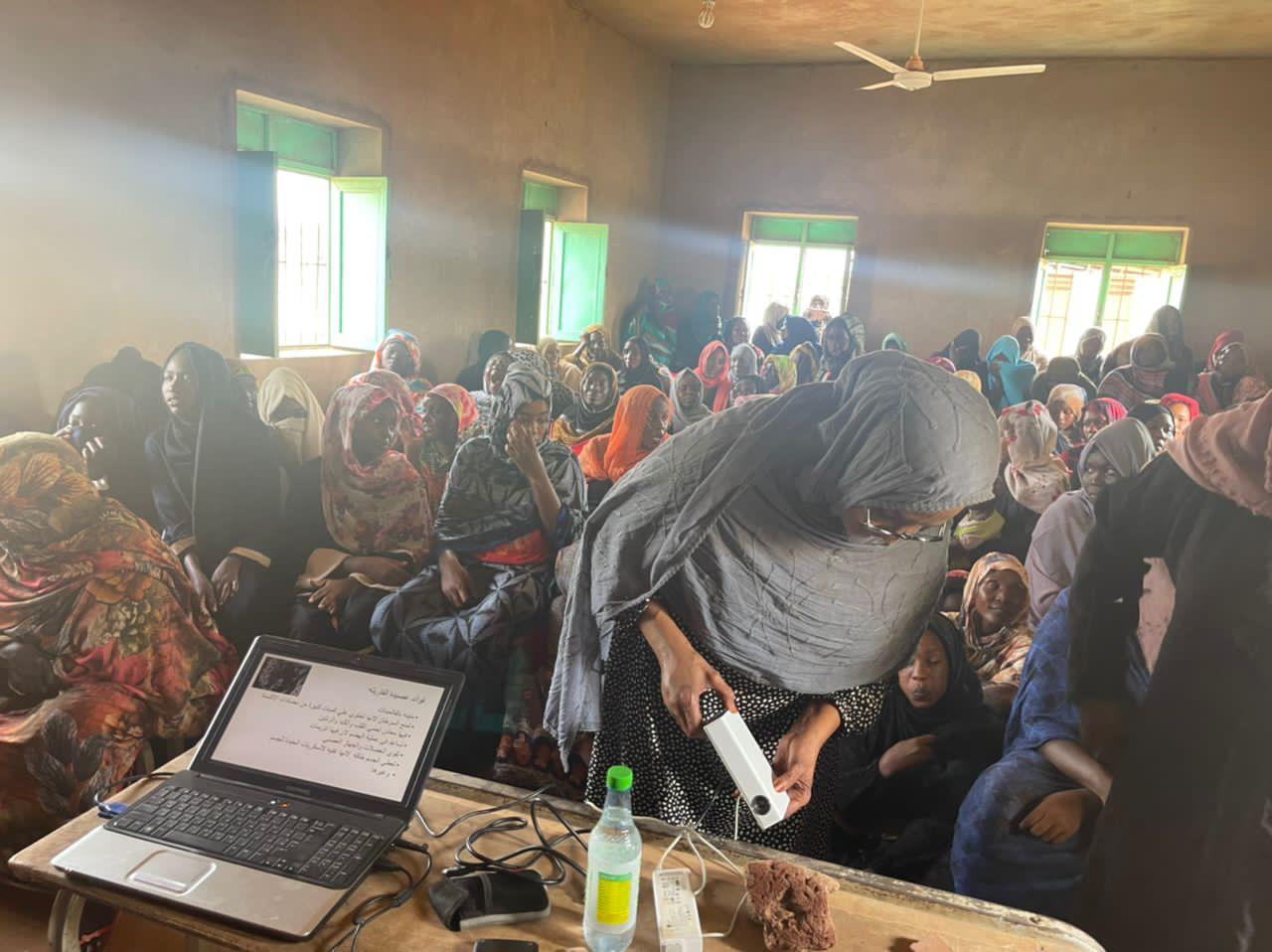 A crowded village hall of women in colourful headscarves; a facilitator leans over to hand out a small device while a laptop on the table shows Arabic slides.