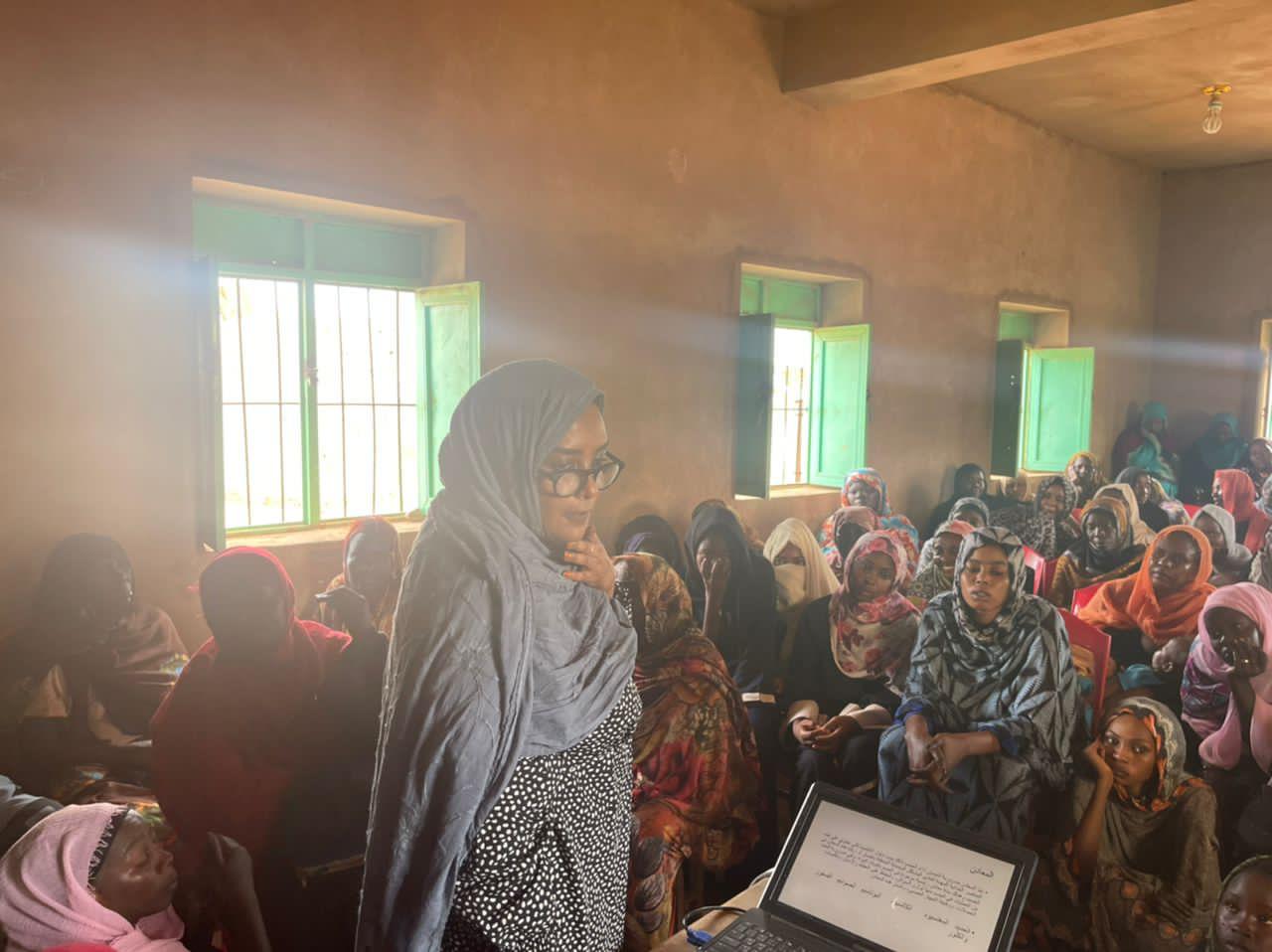 Dr. Nada Alassad, in a grey headscarf and glasses, speaks to a full room of women seated on plastic chairs in a sunlit village hall with green-shuttered windows; a laptop on the table shows slides in Arabic.