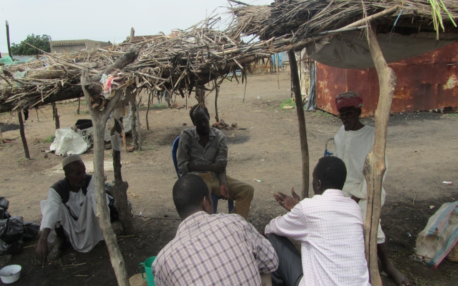 An EBSSA field team in conversation with three community members seated under a shade shelter built from wooden poles and dried branches.