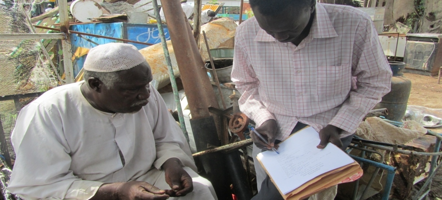 An EBSSA enumerator stands with a clipboard and pen, recording answers while an older man in a white jalabiya and prayer cap speaks to him in a workshop yard.