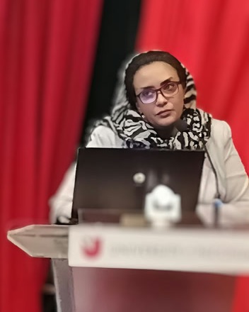 Dr. Nada Alassad, wearing glasses and a patterned headscarf, speaks from a lectern with a laptop in front of her.