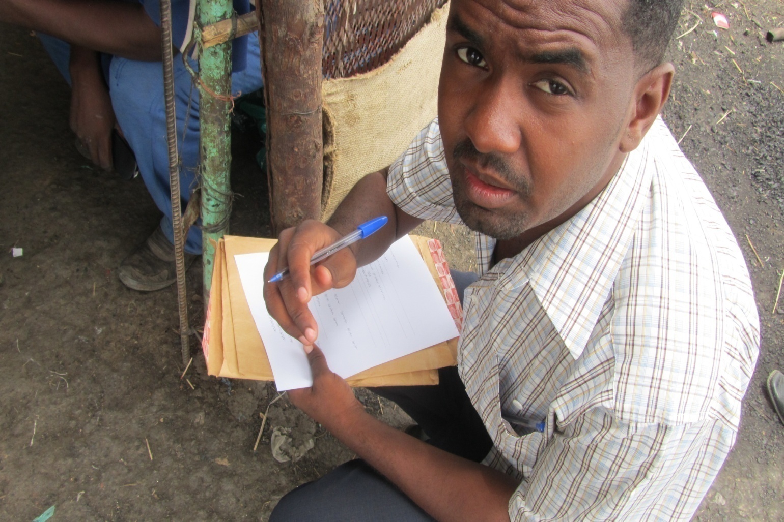 A young EBSSA field researcher crouches with a paper questionnaire on a stack of envelopes, pen in hand, looking up at the camera.