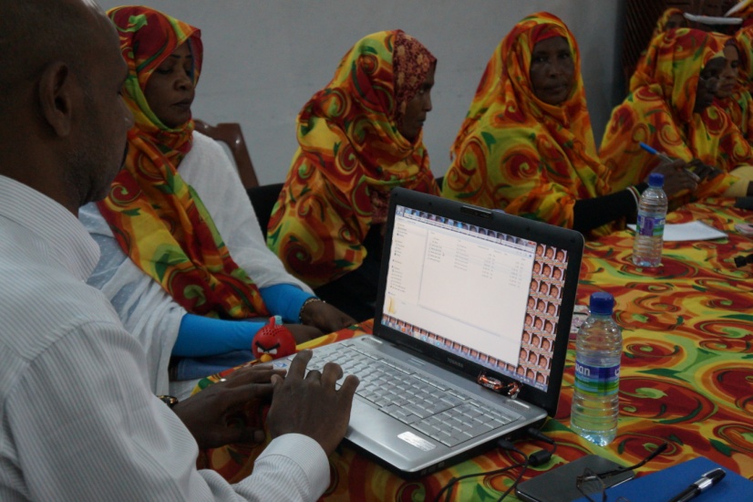 Women in brightly patterned saffron and red tobes sit around a table at a workshop while a facilitator enters data on a laptop.